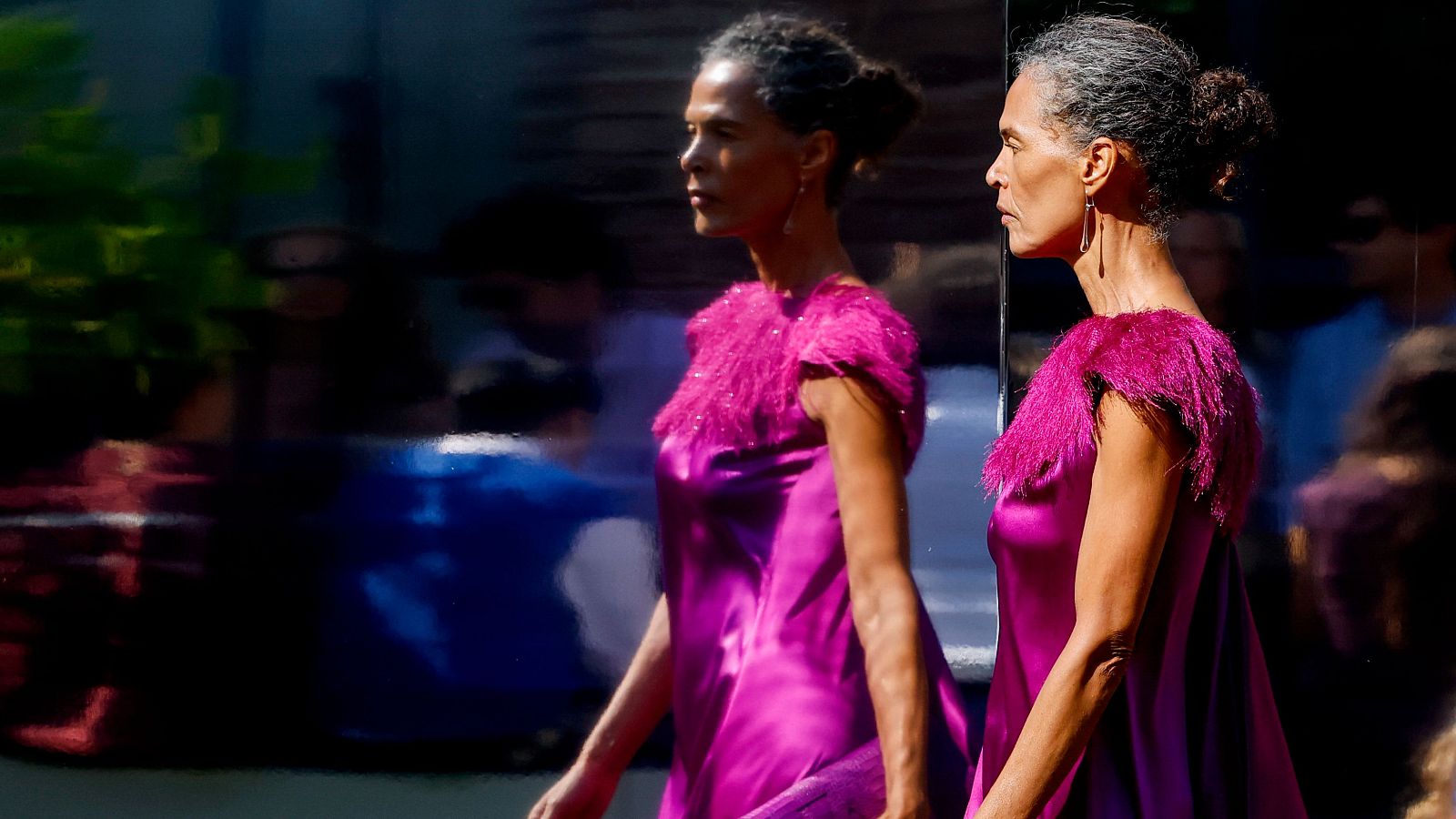 Mujer de perfil con vestido fucsia satinado y hombreras de plumas, reflejada en una superficie brillante. Fondo oscuro y desenfocado.