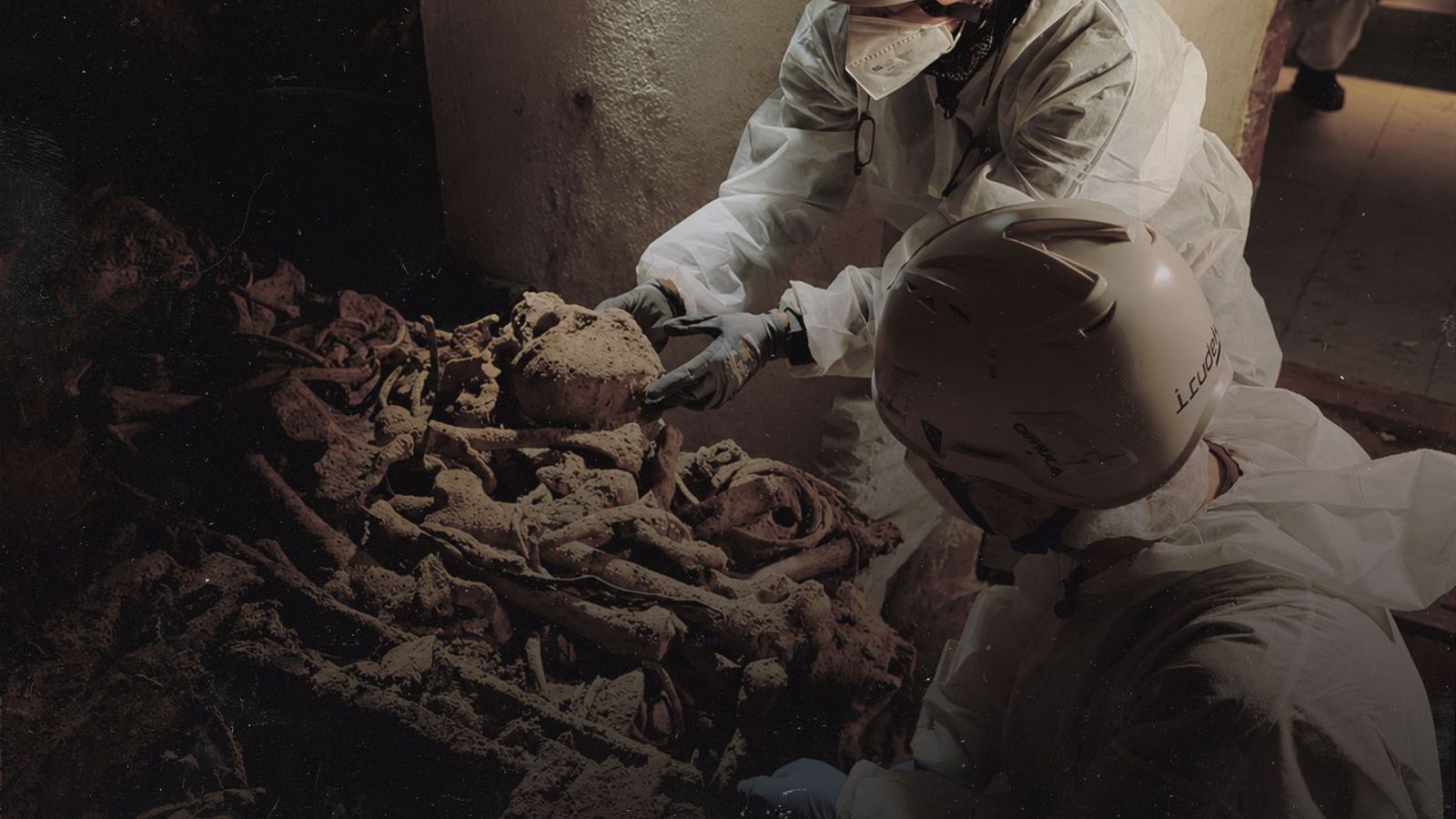 Dos individuos con trajes blancos y cascos, uno con 'I. Trudg' visible, analizan una gran cantidad de huesos y cráneos humanos en un área sombría.