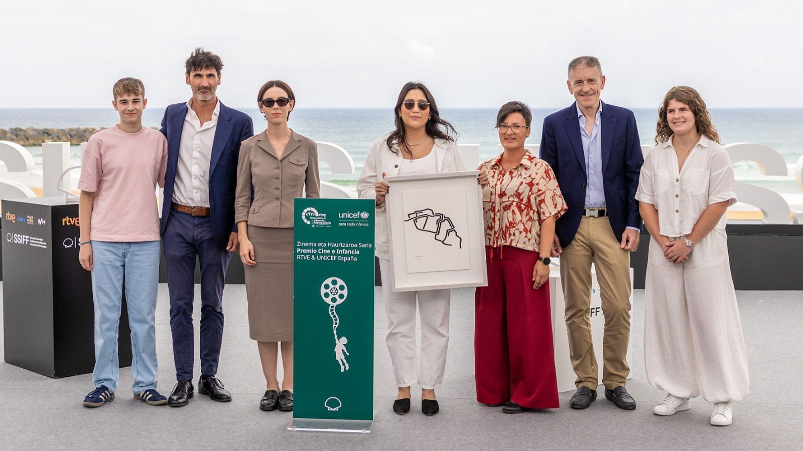 Siete personas posan frente a las letras "SSIFF" y el mar. Antonio de la Torre está presente. Un banner con logotipos de UNICEF y RTVE, y textos en euskera y español, se ve en la imagen.