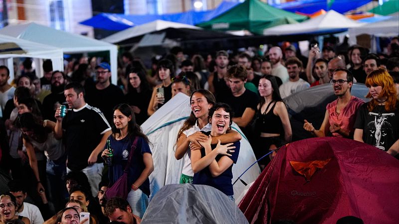 Dos jóvenes se abrazan y ríen alegremente en medio de una concurrida acampada nocturna rodeadas de tiendas de campaña y otros jóvenes.