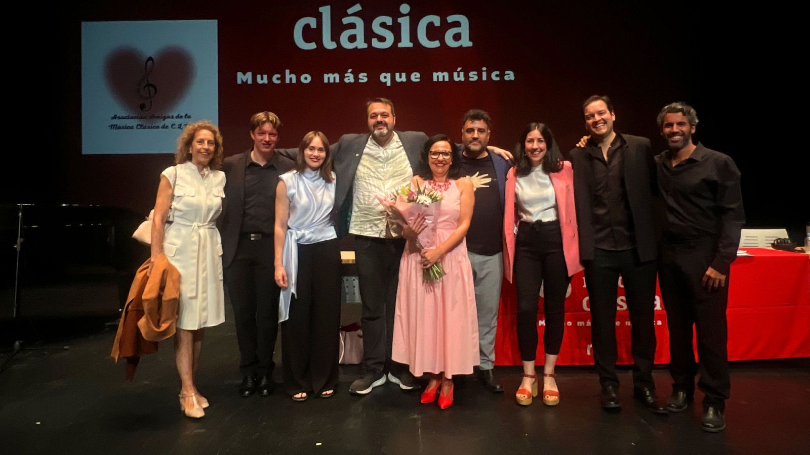 Nueve personas, incluido el periodista Martín Llade, posan en un escenario ante una pantalla roja con el logo de la Asociación de Amigos de la Música Clásica de Castilla-La Mancha.