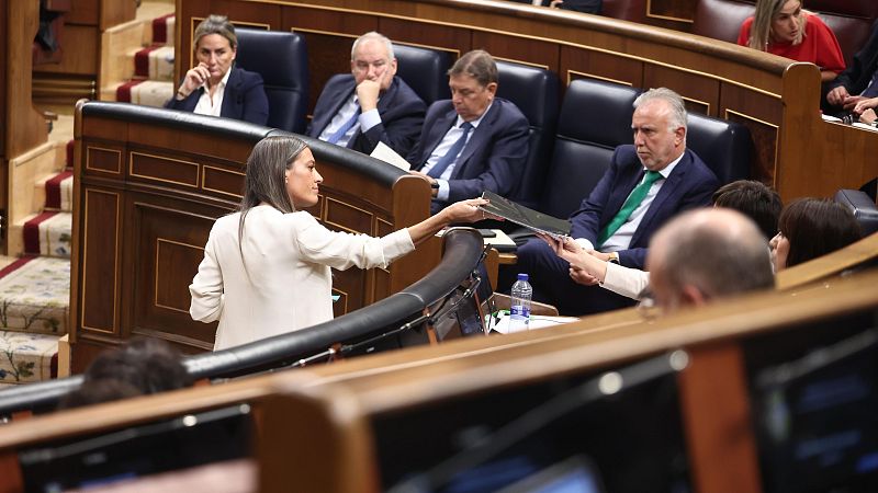 Una mujer de pie con chaqueta marfil entrega una carpeta negra a una ministra sentada, mientras otros ministros observan la escena en el hemiciclo.