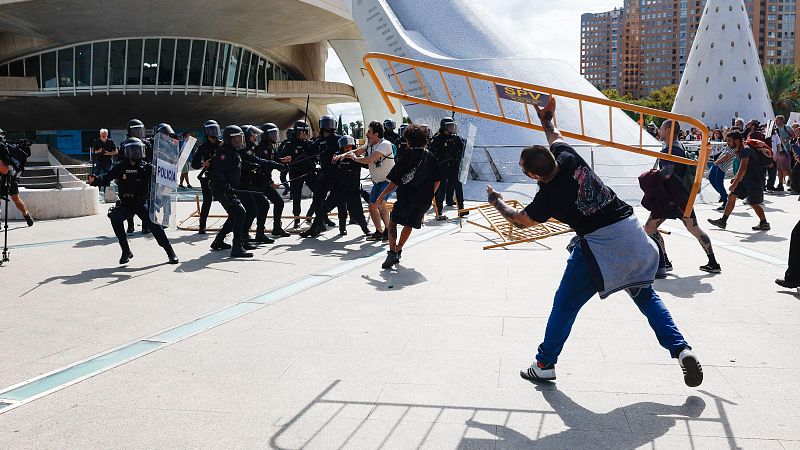 Agentes antidisturbios de la Policía Nacional y manifestantes se enfrentan en una explanada, con un joven levantando una valla.