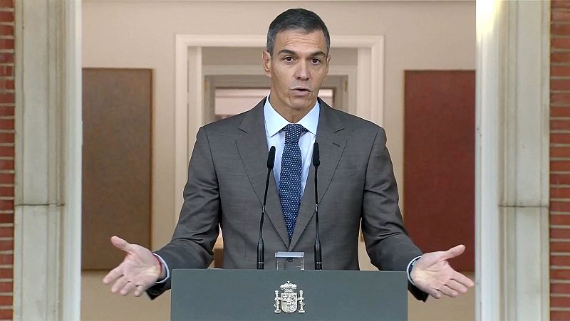 Pedro Sánchez comparece serio ante un atril con el escudo de España, haciendo un gesto con las manos mientras viste traje gris y corbata azul.