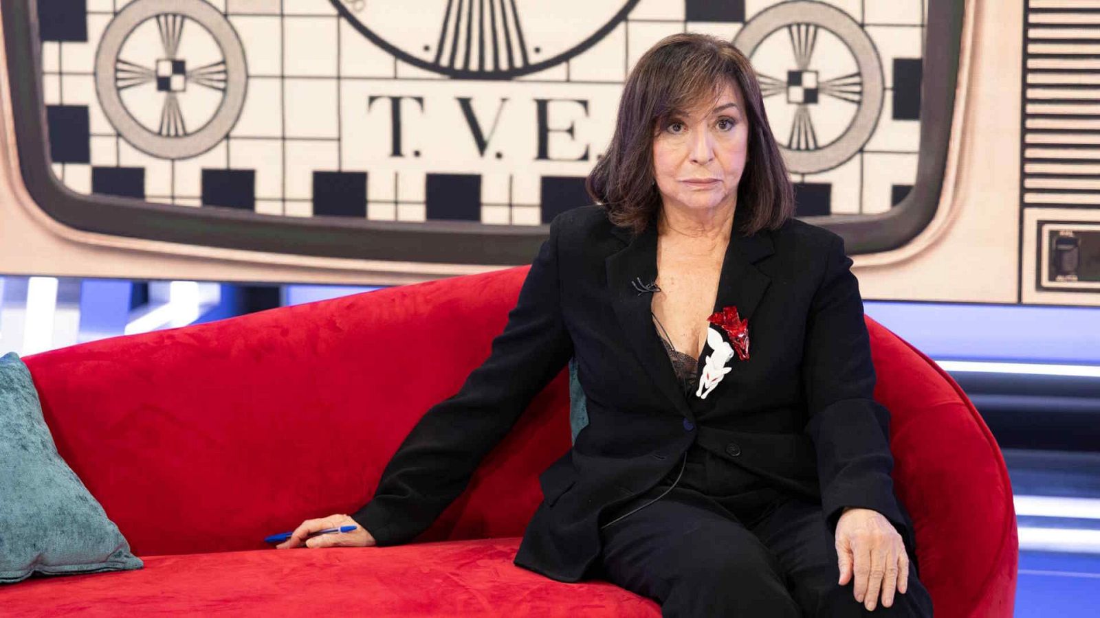 La periodista Ana Blanco, con traje negro y broche, está sentada en un sofá rojo ante un panel retroiluminado con la clásica carta de ajuste de televisión.