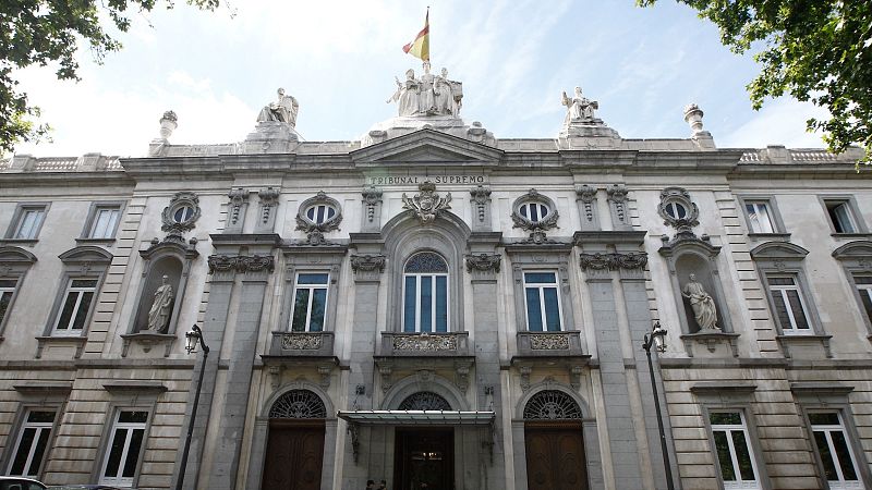 Plano contrapicado de la fachada clásica de piedra del Tribunal Supremo en Madrid, con inscripciones, esculturas, balcones y una bandera de España.