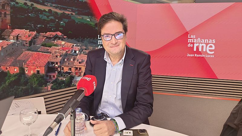 El periodista Íñigo Alfonso sonríe ante un micrófono de radio en un estudio con paneles decorativos y el logotipo del programa de RNE.